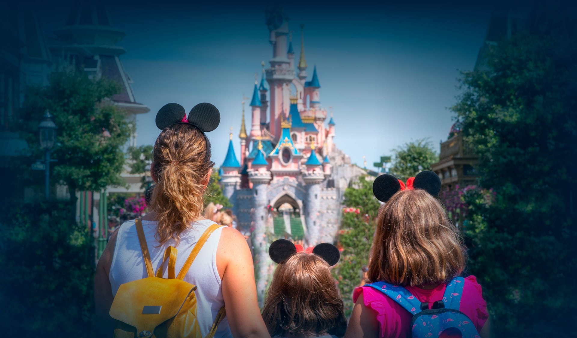 Tres personas con orejas de Mickey Mouse están de espaldas a la cámara, frente a un castillo rosa en un parque temático.