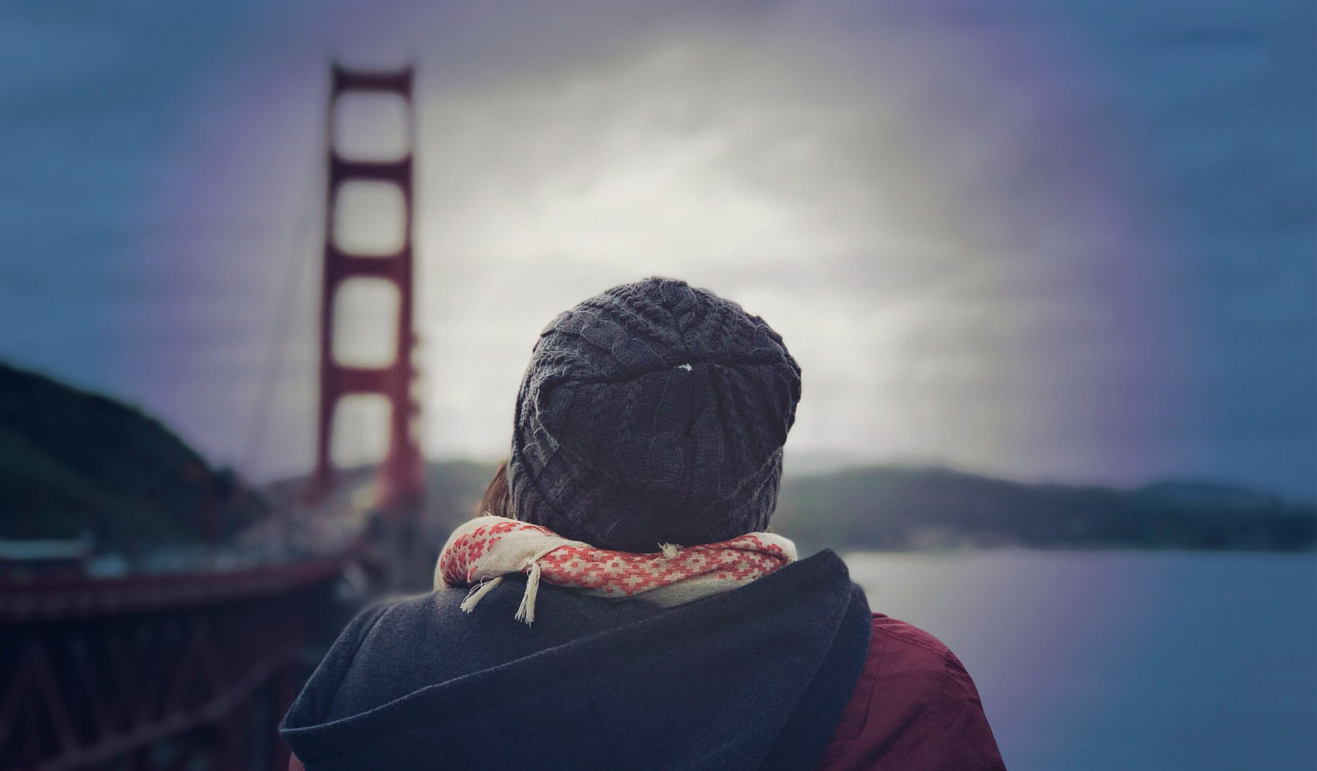 Persona con gorro de punto y bufanda frente al puente Golden Gate en un día nublado.