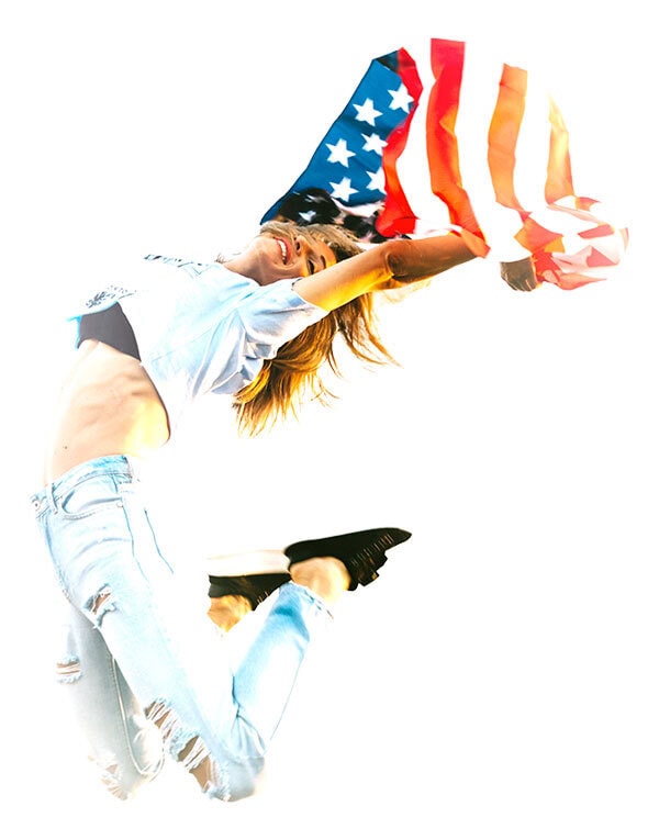 Una persona con vaqueros rotos y camisa clara salta en el aire sujetando una bandera de Estados Unidos.